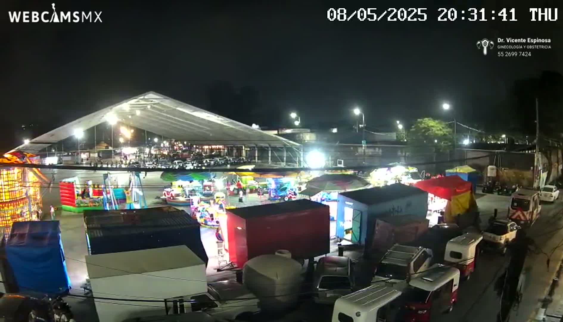 A nighttime aerial view shows a fairground with carnival rides and food stalls under a large tent, surrounded by parked vehicles and under a clear, dark sky.
