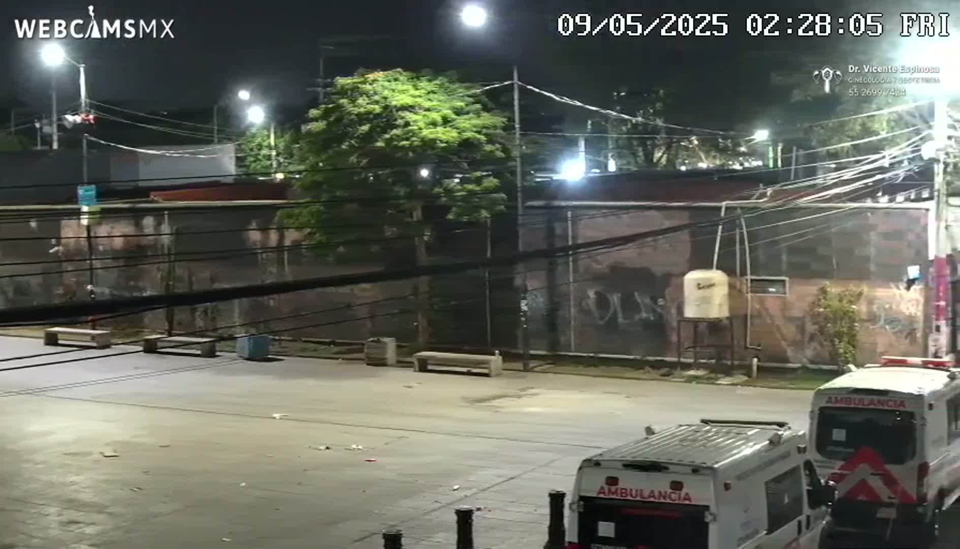 At night, two ambulances are parked in an empty lot next to a graffiti-covered wall, under bright streetlights.