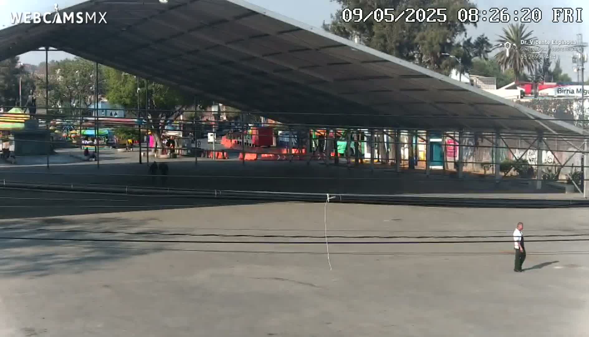 A mostly empty paved area is sheltered by a large metal roof structure, overlooking a park with carnival rides under a sunny sky.