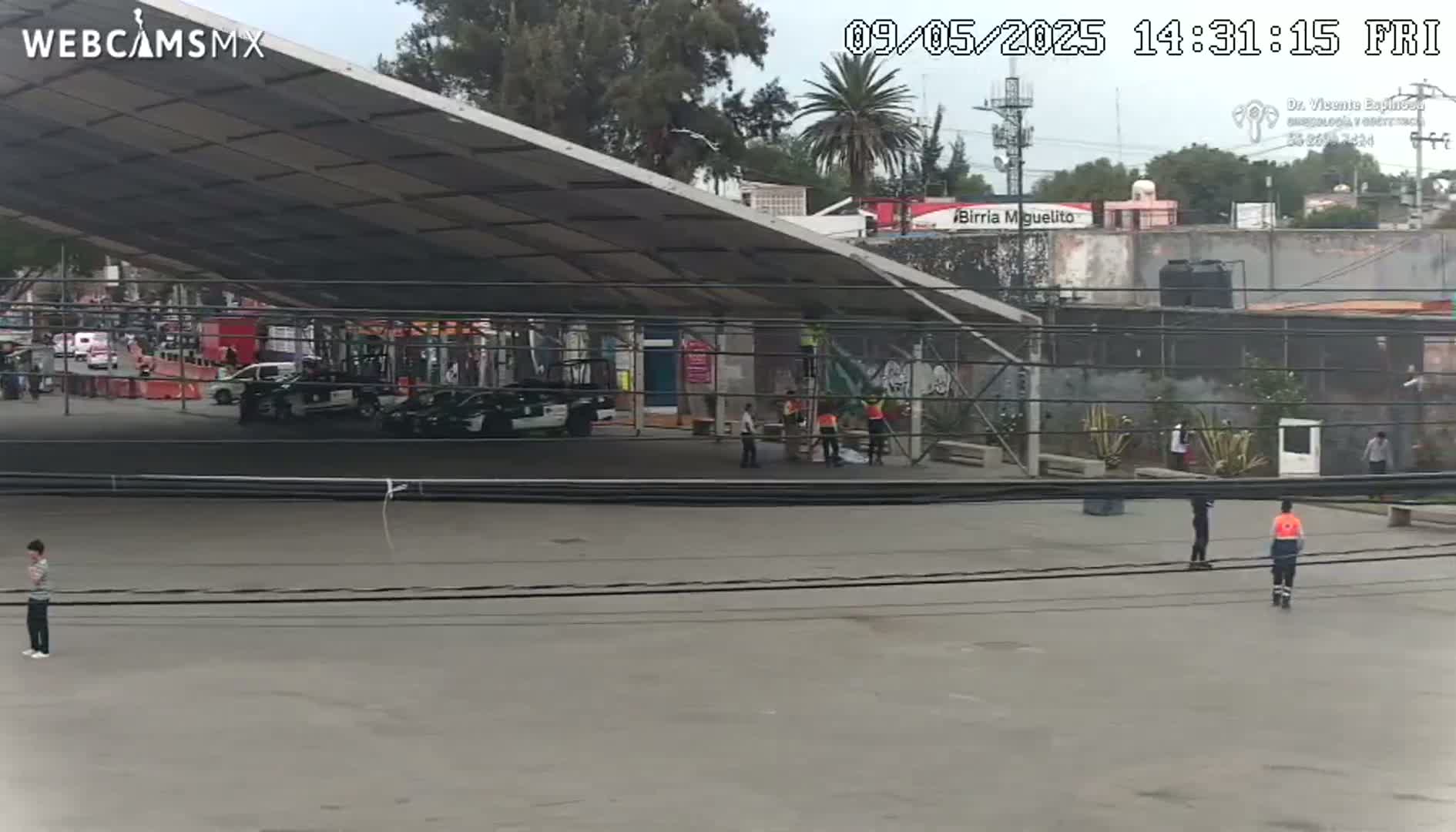 Under a large metal awning, several police cars are parked in a mostly empty paved lot, with a few people standing around and some construction work taking place nearby, under a partly cloudy sky.