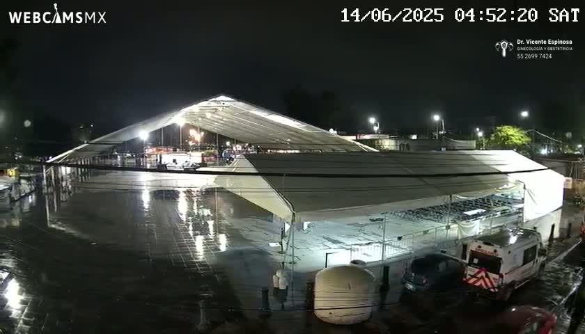 A large, white tent structure sits in a rain-soaked paved area at night, with several vehicles and a few people visible nearby.