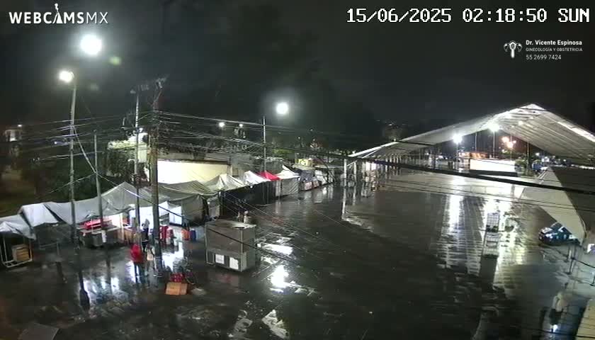 A wet, nighttime scene shows numerous tents set up in a parking lot under a large awning, with streetlights illuminating the area.