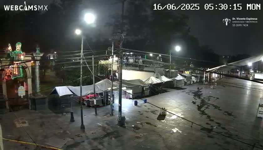 A nighttime, wet street scene shows several tents and temporary structures set up near a building with illuminated religious iconography.