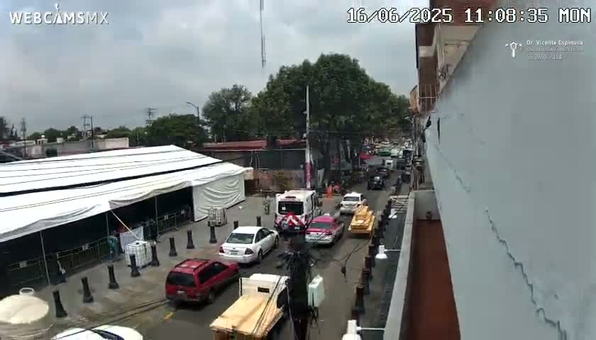 A street scene on an overcast day shows several cars and trucks parked and driving near a large white tent and a building under construction.