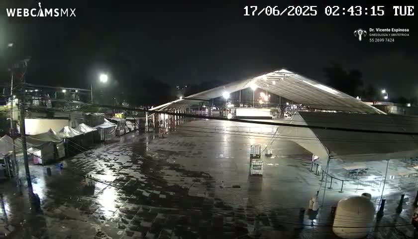 A nighttime view of a large, wet paved area with tents and a large covered structure, under rainy conditions.