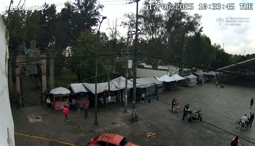 An overcast day reveals a paved plaza with numerous market stalls under gray canopies, surrounded by trees and a stone archway, with people walking about and a few vehicles present.