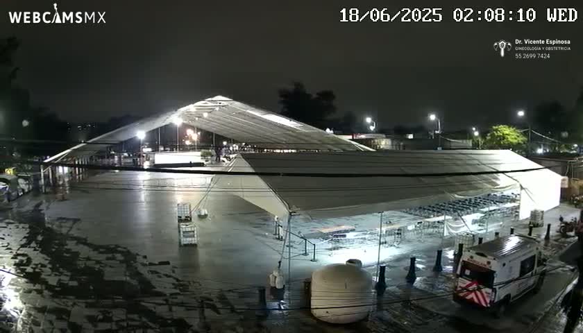A large, illuminated outdoor structure, possibly a temporary market or event space, is wet from recent rain under a dark, nighttime sky.
