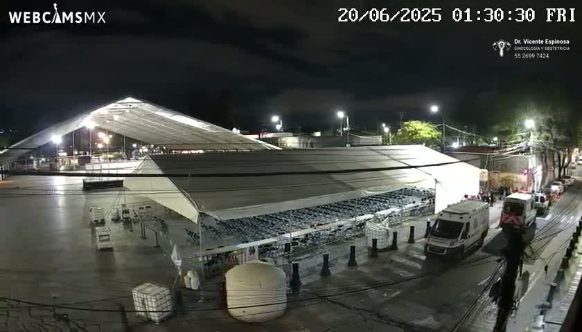 A large, white, covered structure, possibly a tent, sits in a mostly empty parking lot at night, with a few vehicles and people visible in the distance.