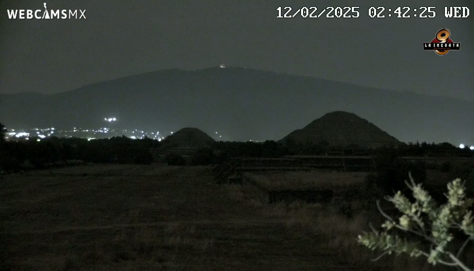 At night, under a dark sky, several dark pyramids sit on a flat, dark plain, with a distant mountain range in the background and city lights visible on the horizon.