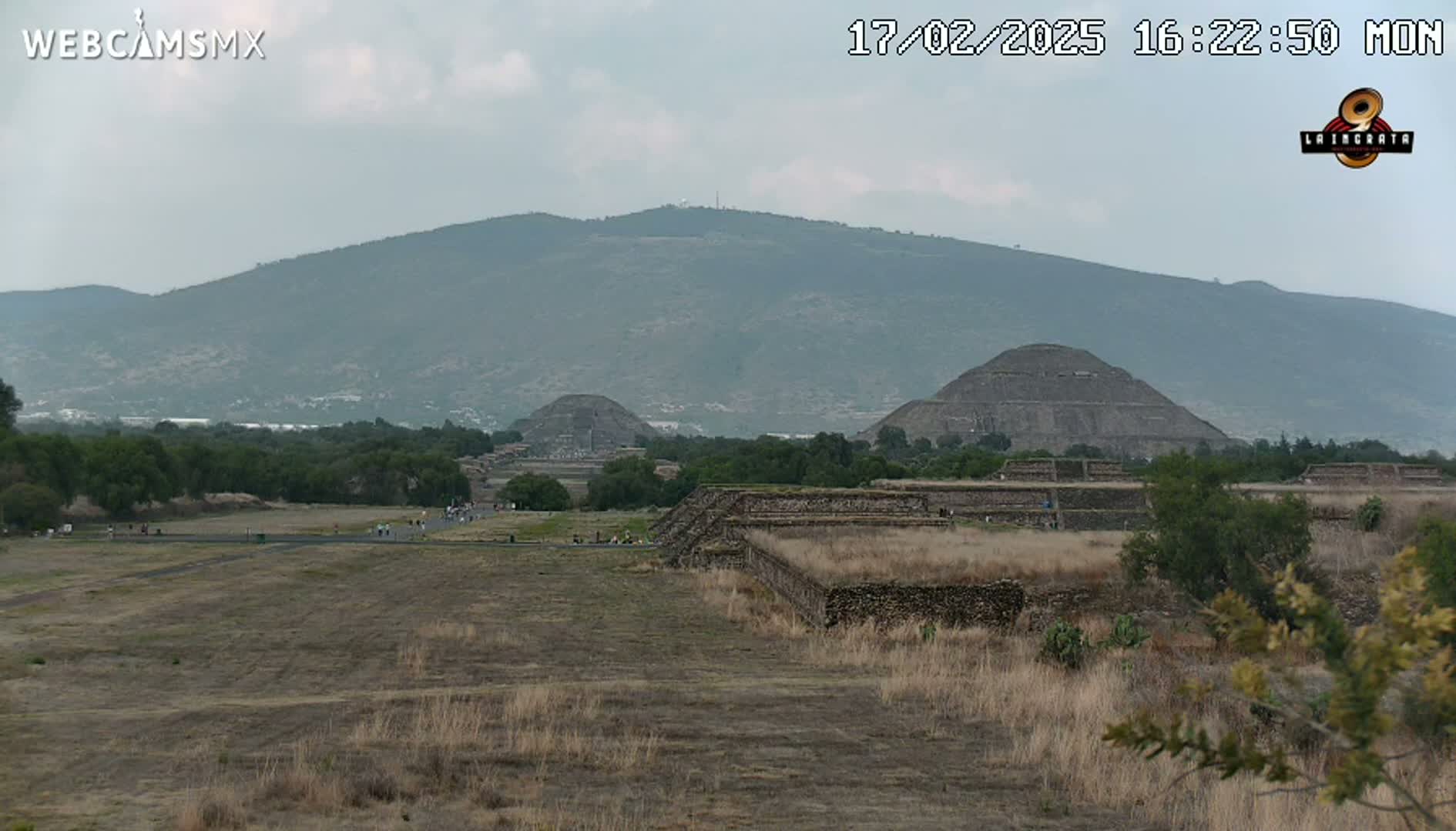 A wide, flat expanse of dry, brown grass leads to several ancient stone pyramids and a large mountain under an overcast sky.