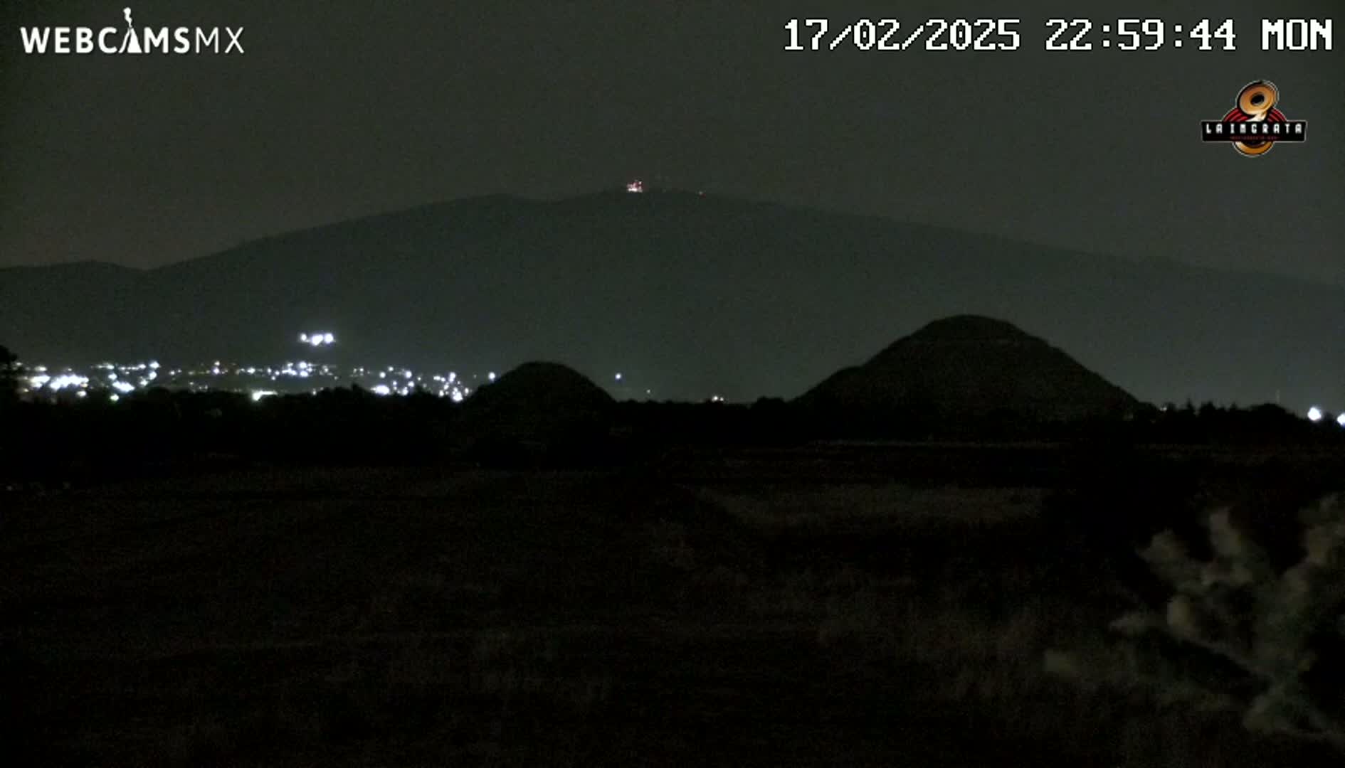 A nighttime view shows dark, low hills in the foreground, with a larger, darker hill and a distant city's lights in the background, under a clear, dark sky.