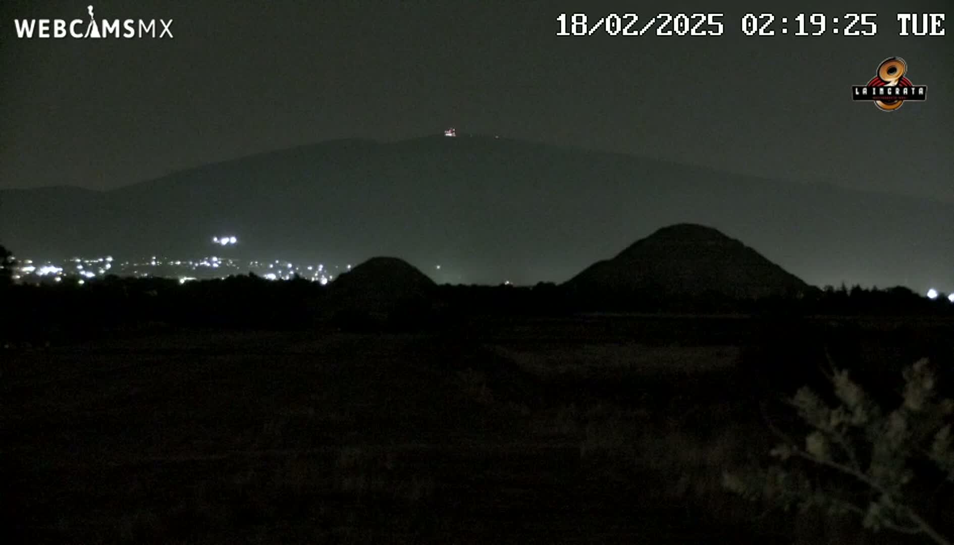 At night, a dark landscape shows two hills in the foreground and a mountain range in the background, with a few distant lights visible.