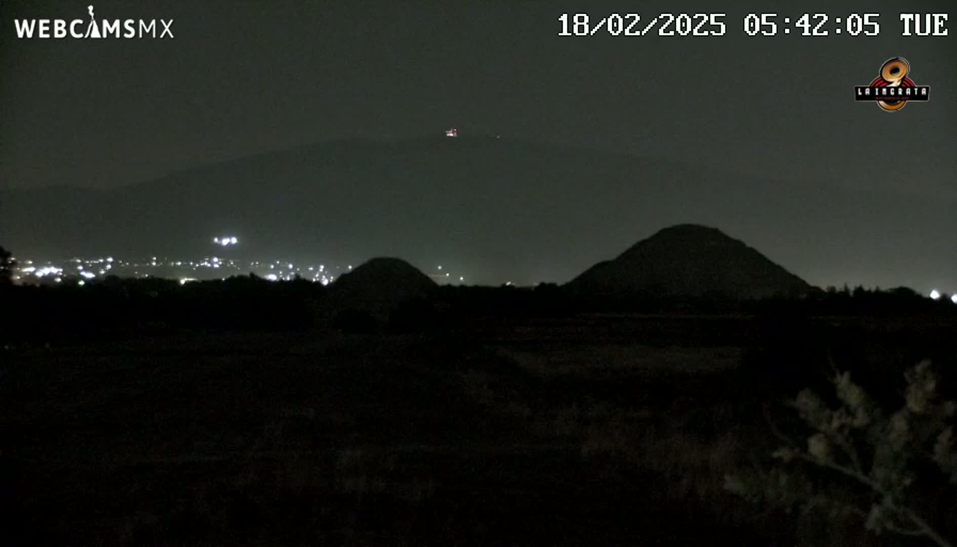 At night, a dark landscape shows two hills in the mid-ground and a mountain range in the background, all silhouetted against a dark sky with a few distant lights visible.