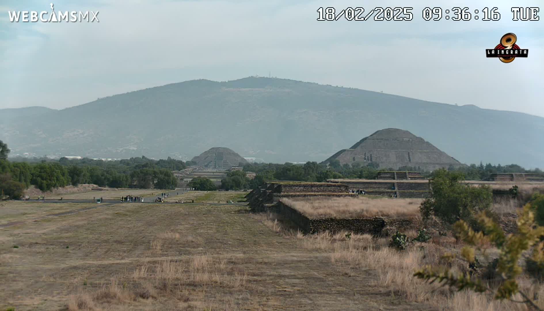 A hazy, long shot shows several ancient pyramids and stone structures on a dry, grassy plain, with a large mountain in the background.