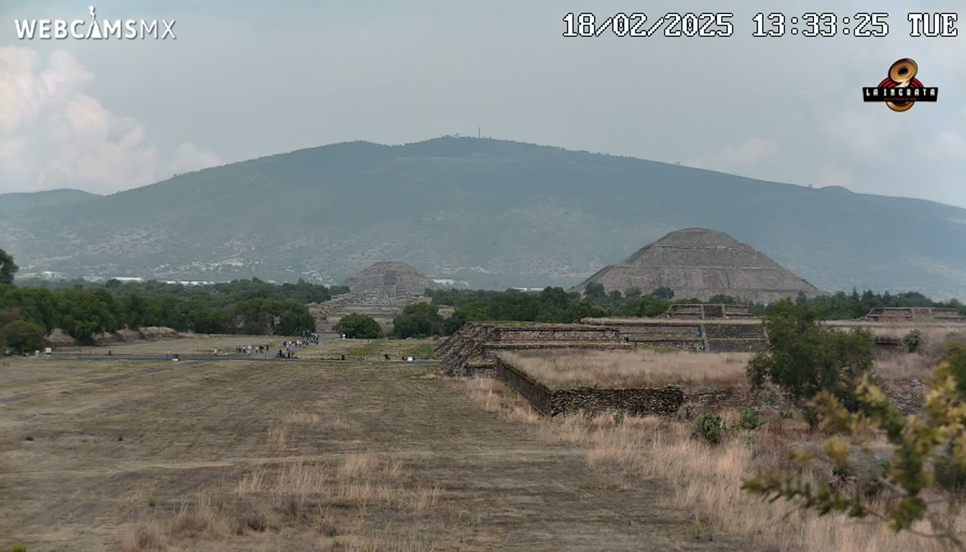 A hazy view of Teotihuacan pyramids and ruins stretches across a dry, brown landscape, with a mountain range in the background under a cloudy sky.