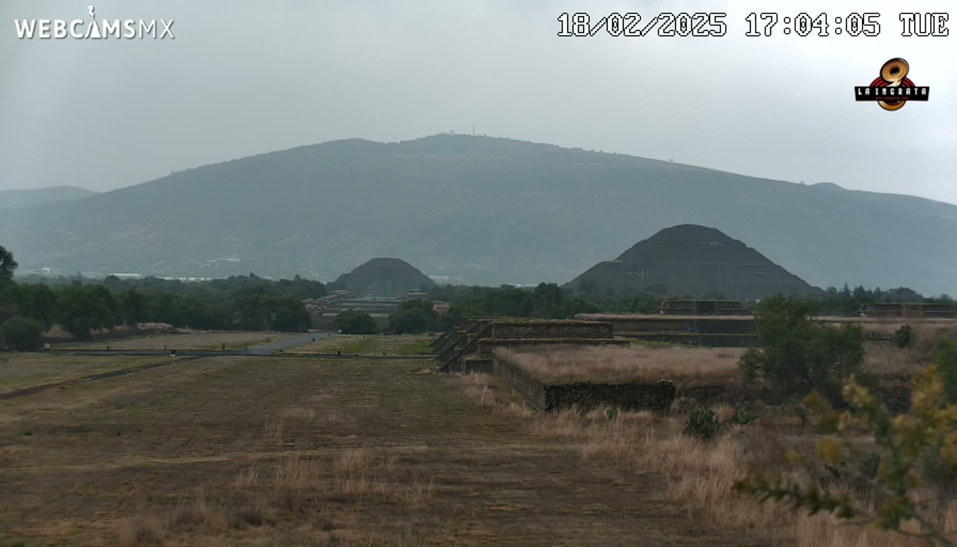 A hazy view of ancient pyramids and stone structures on a flat, dry landscape, with a mountain range in the background under an overcast sky.