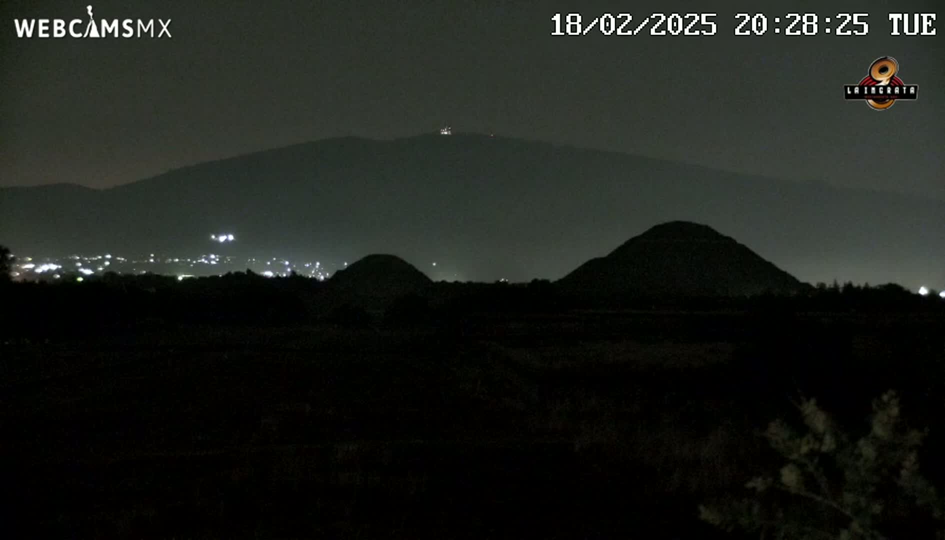 At night, under a dark sky, two dark, rounded hills are silhouetted against a hazy mountain range in the distance, with a few faint lights visible in a town below.