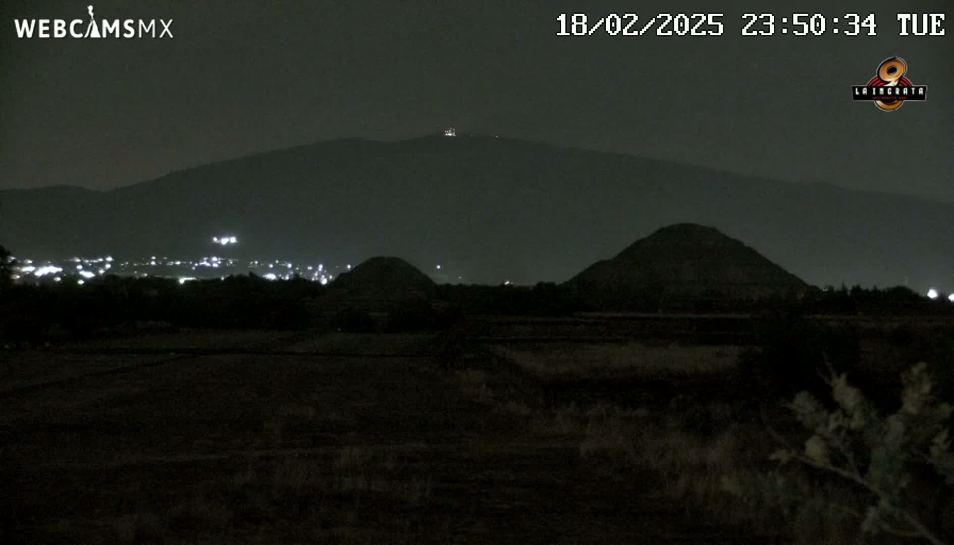 A nighttime view of a dark landscape featuring two low hills and a distant mountain range under a clear, dark sky.