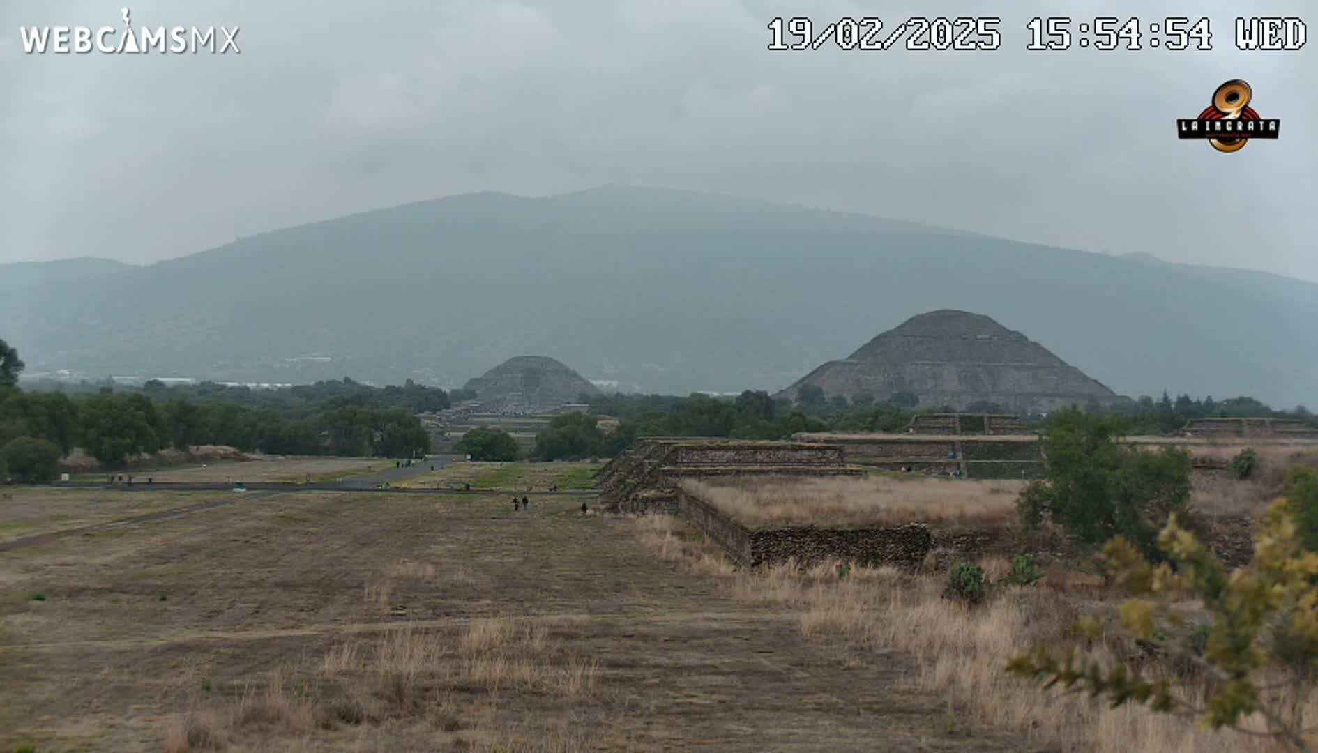 A hazy view of several ancient stone pyramids and surrounding ruins, set against a backdrop of mountains under a cloudy sky.