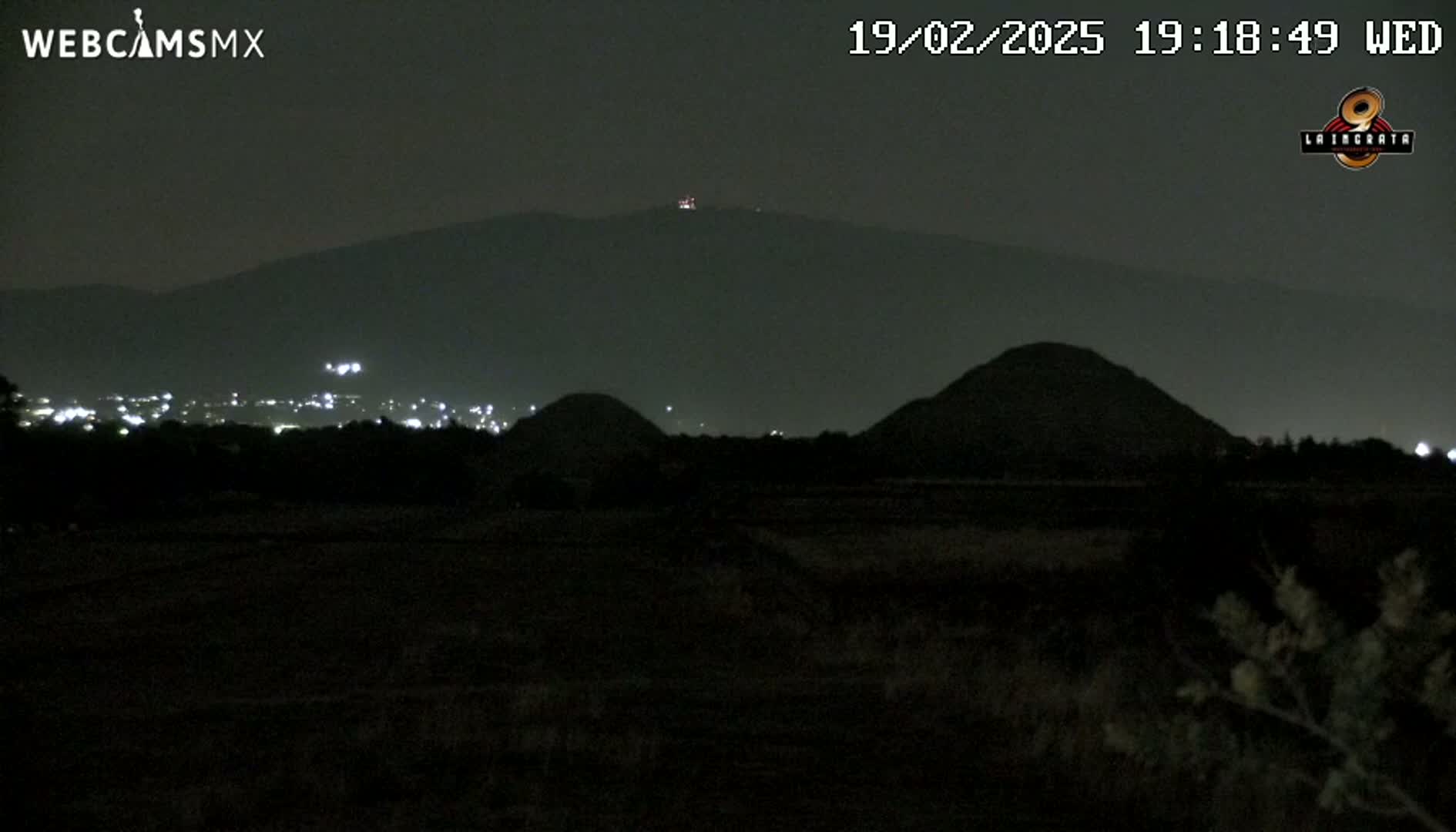 A nighttime, dark view of several hills and a distant city with faint lights, under a clear, dark sky.