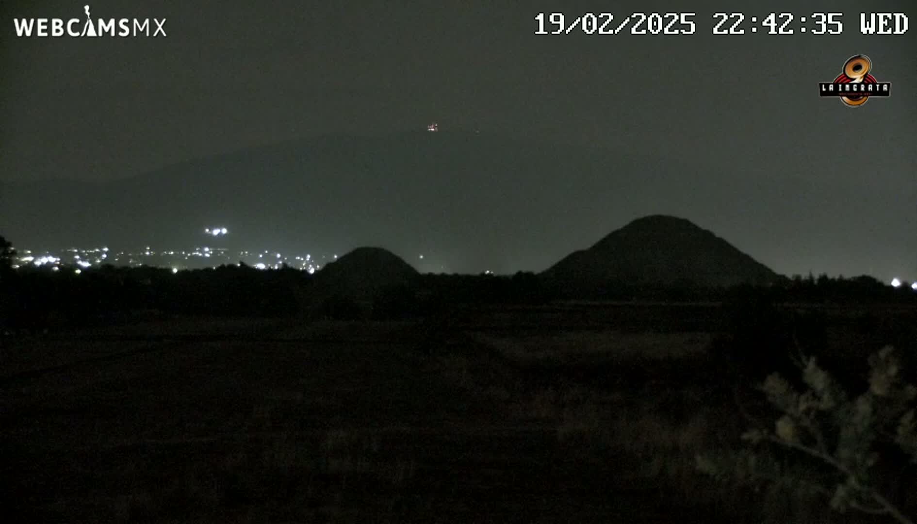 At night, a dark landscape shows two hills in the distance, with a city's lights faintly visible beyond them.