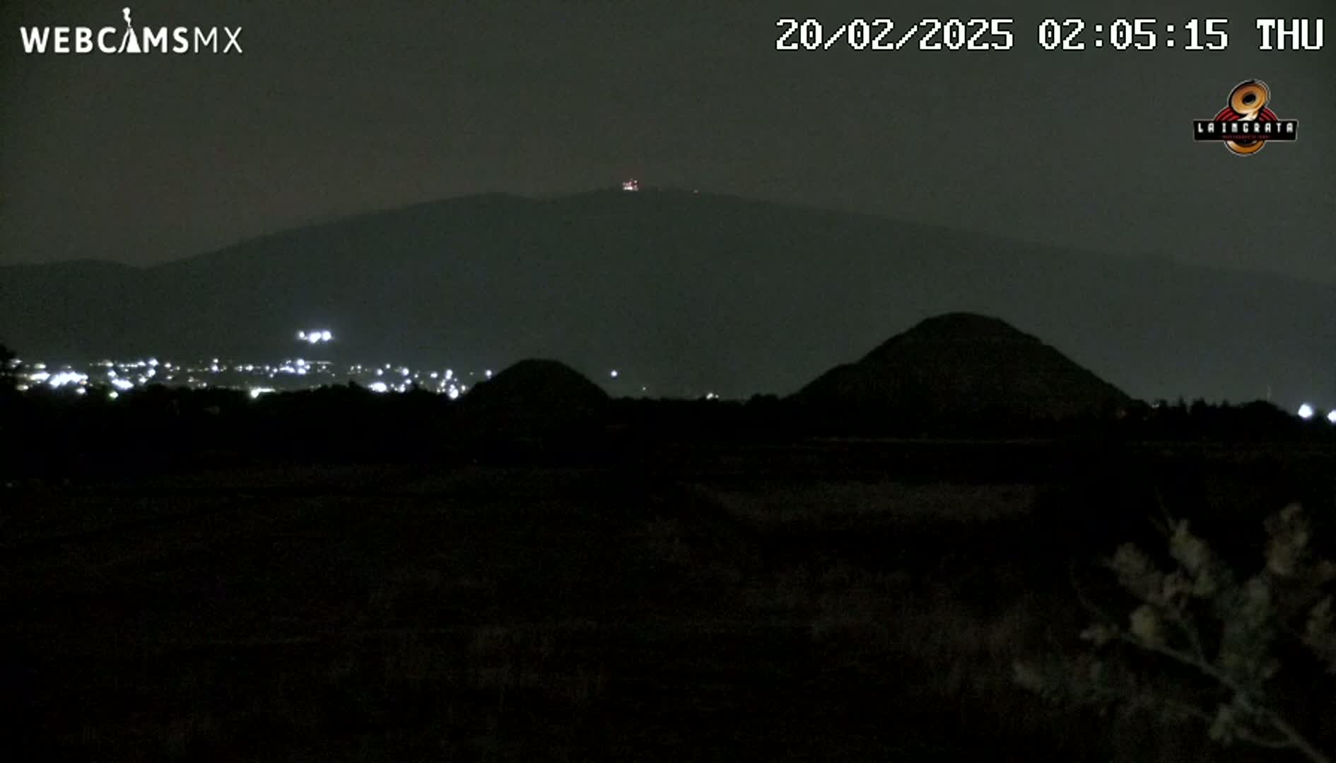 At night, a dark landscape shows several low hills silhouetted against a distant mountain range under a clear sky.