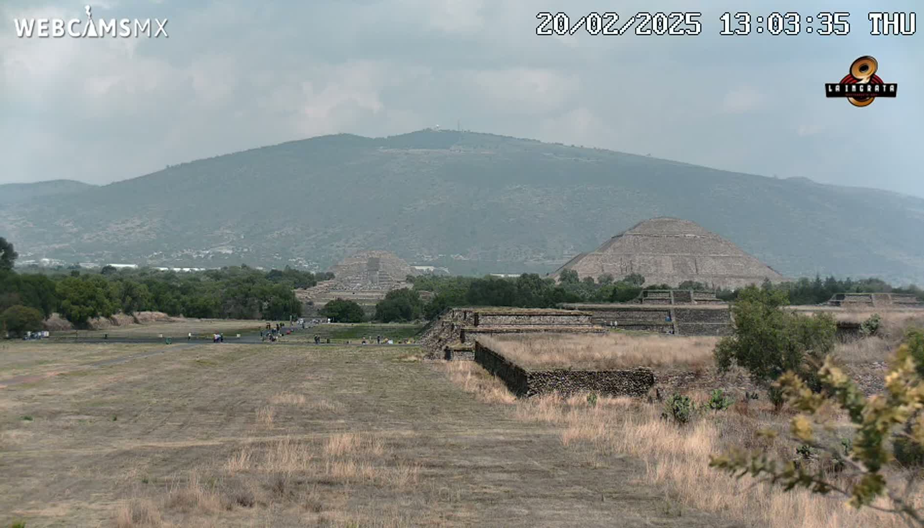 A wide, flat, dry field with low-lying vegetation stretches towards ancient stone pyramids and a hazy mountain range under a cloudy sky.