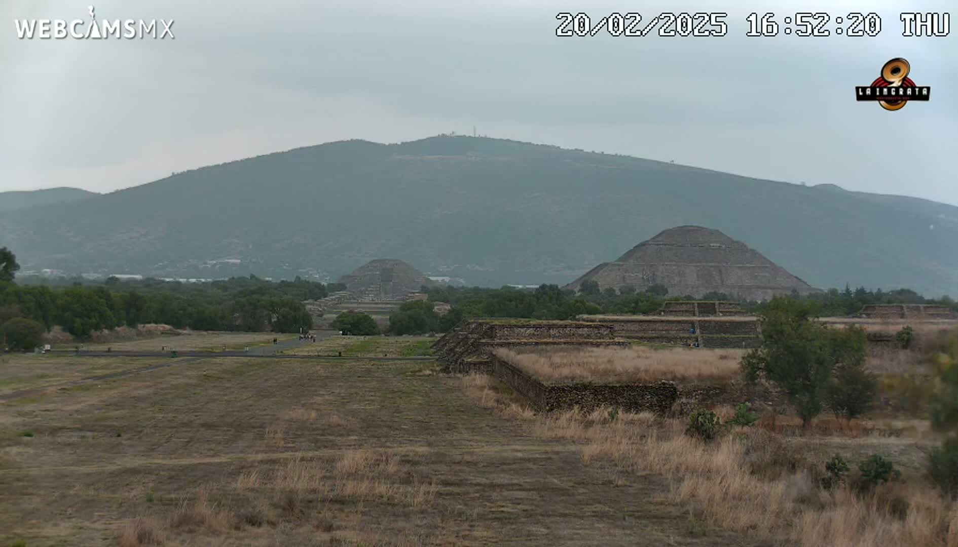 A view of several ancient stone pyramids and structures in a dry, grassy field under an overcast sky, with mountains in the background.
