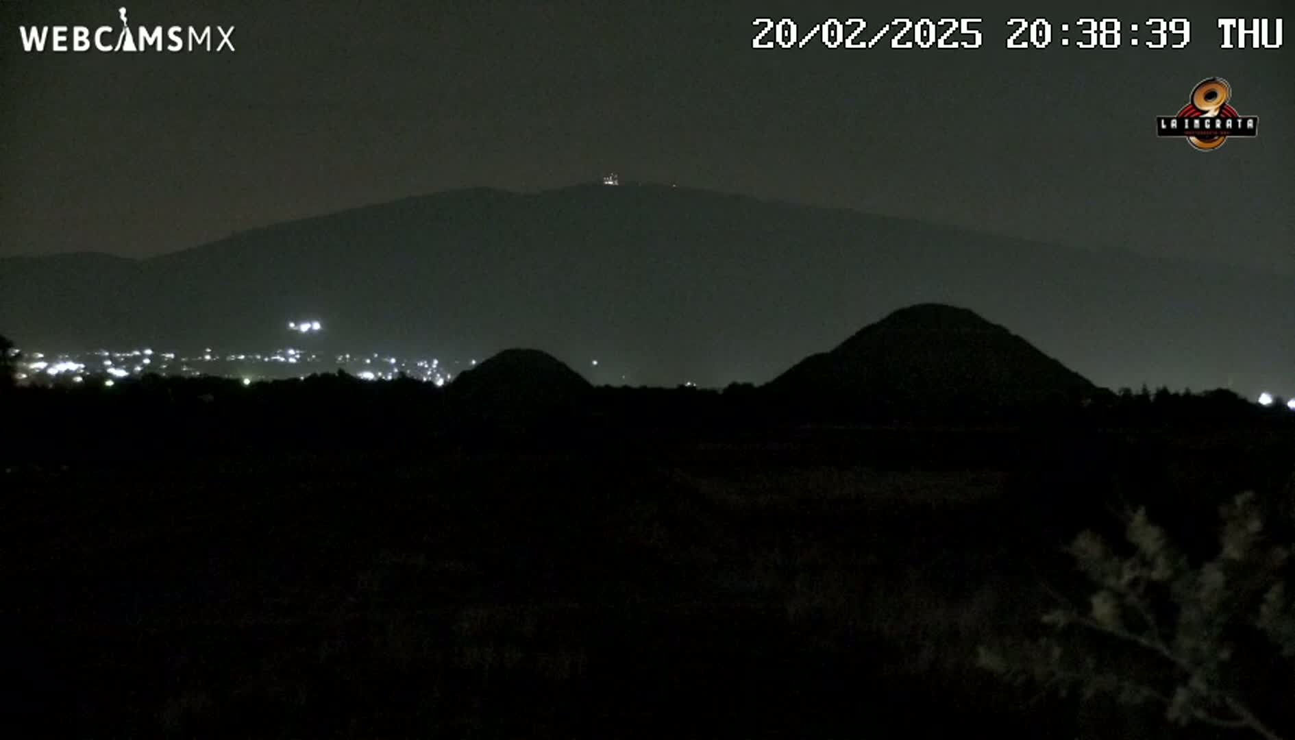 A nighttime view of dark hills and a distant city illuminated by faint lights under a clear, dark sky.