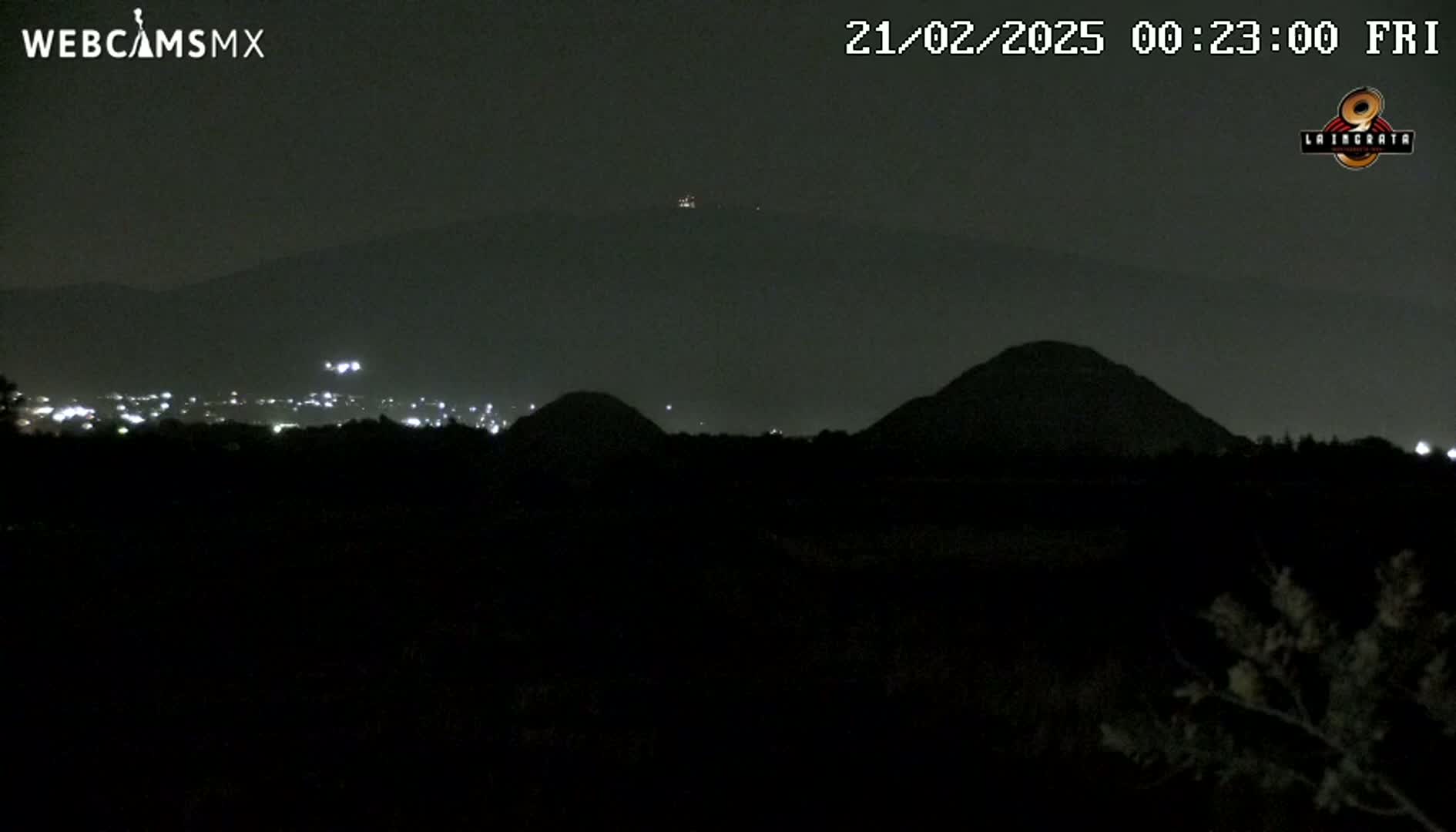 A nighttime view of dark hills and a distant city illuminated by faint lights under a clear, dark sky.