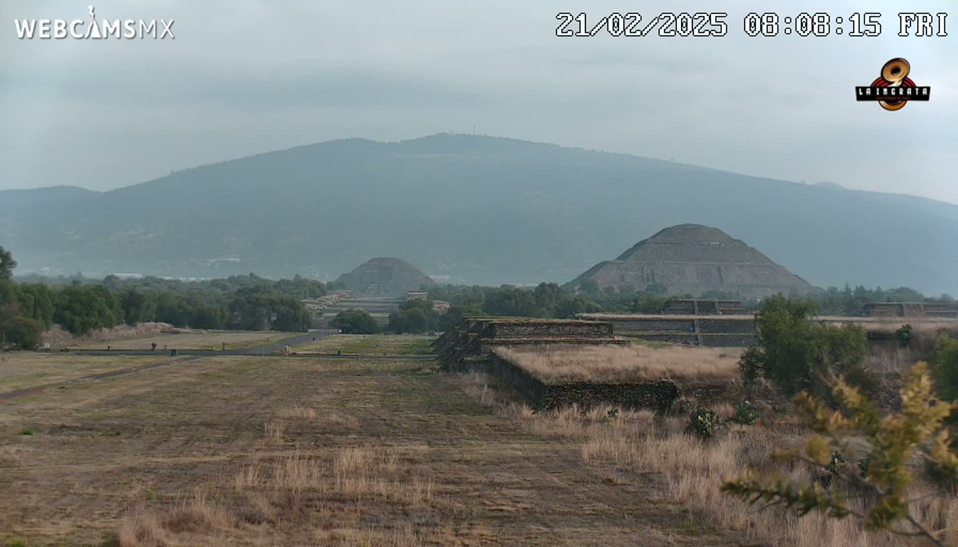 A hazy view of ancient stone pyramids and structures set against a backdrop of a distant mountain range under a cloudy sky.