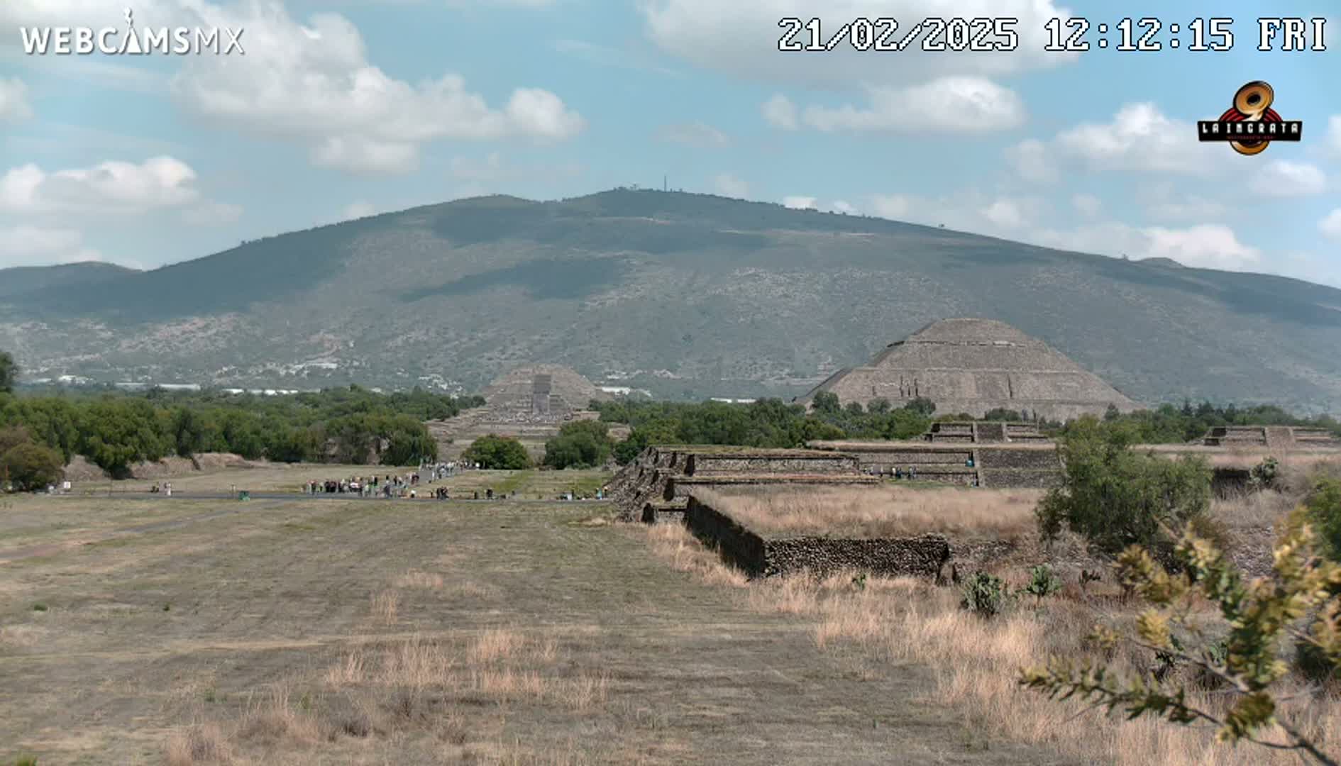 A partly cloudy day reveals several ancient stone pyramids and structures situated in a dry, grassy field, with a large mountain in the background.