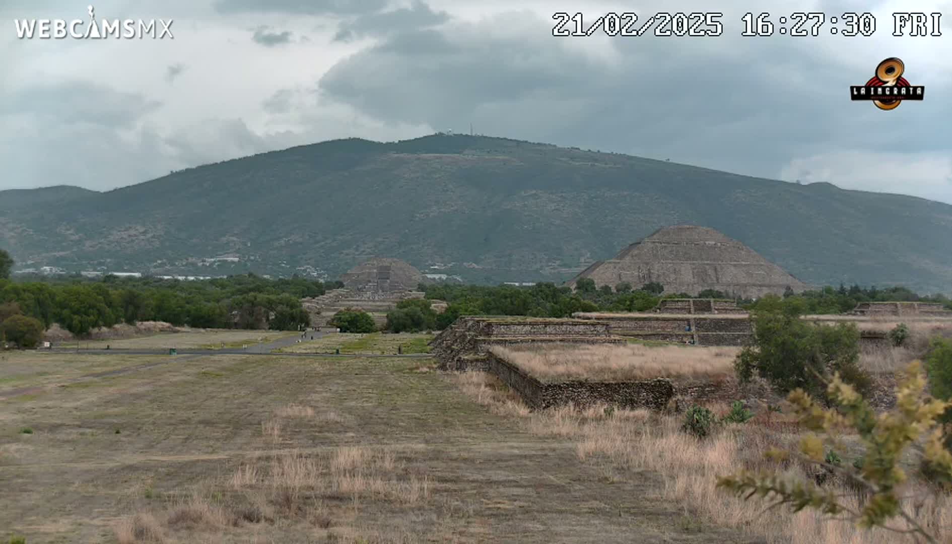 A view of ancient stone pyramids and other ruins set against a backdrop of a mountain under a cloudy sky.