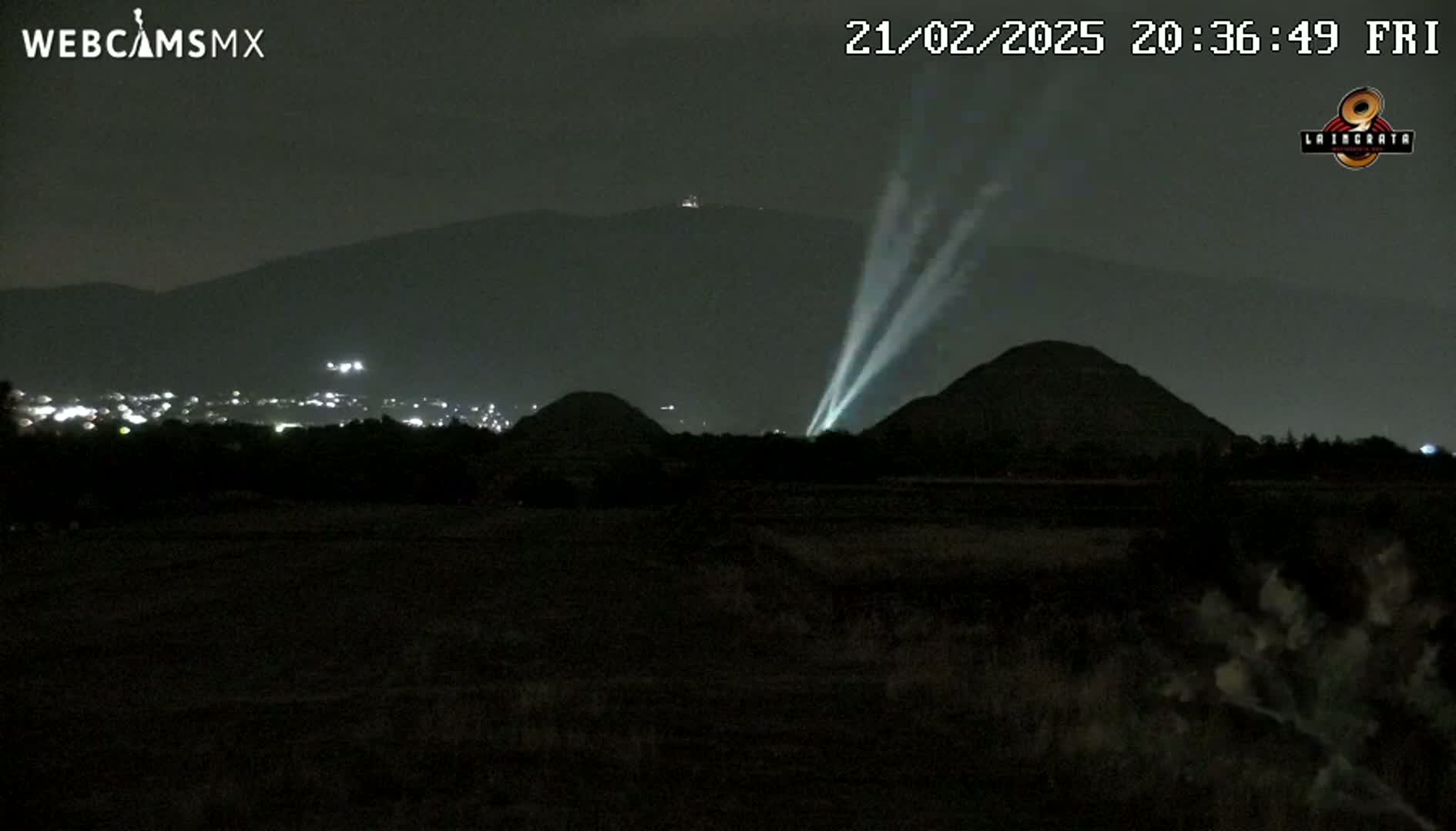 At night, spotlights illuminate two hills in the foreground, with a city and mountains visible in the distance.