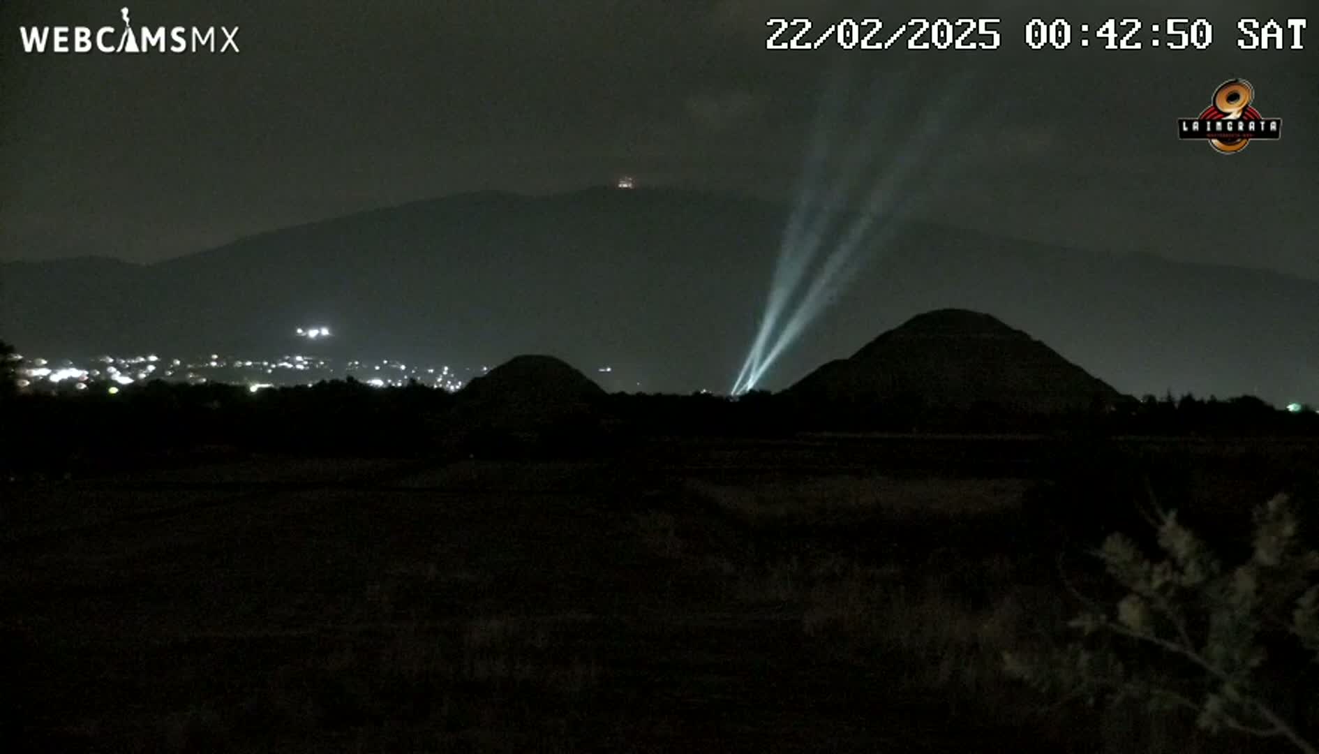 At night, bright beams of light illuminate two dark mounds in front of a distant mountain range under a clear, dark sky.