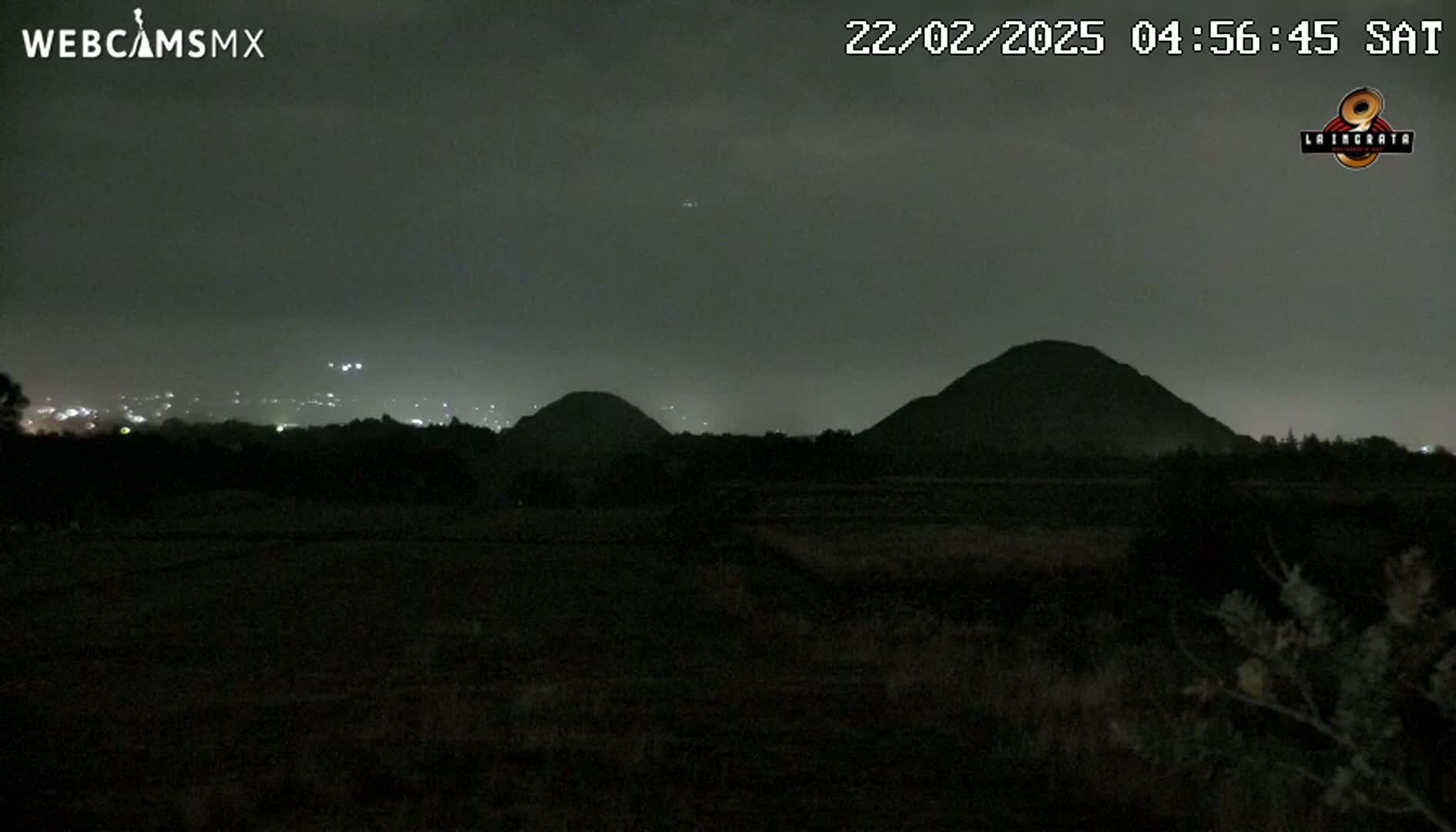 At night, under a dark sky, two large, dark hills are silhouetted against a dimly lit cityscape in the distance.