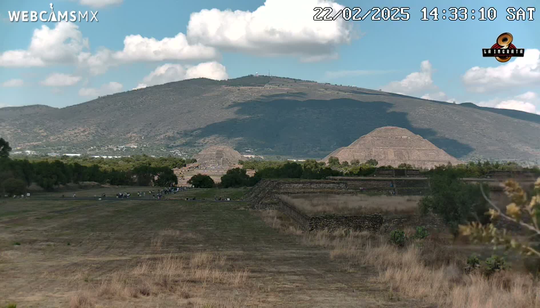 Under a partly cloudy sky, several ancient stone pyramids and structures sit on a large, dry, grassy plain with a mountain in the background.