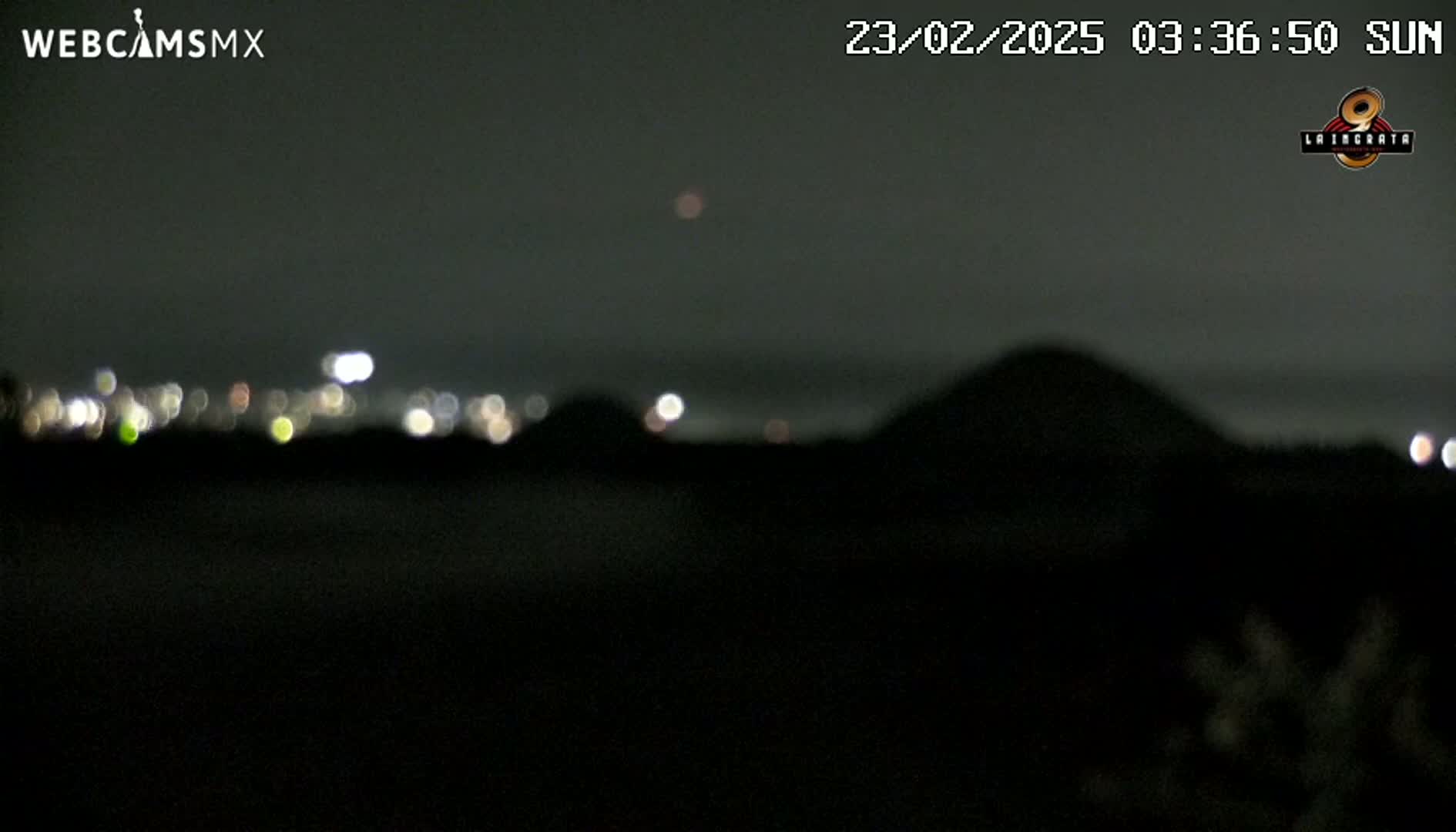 A blurry, nighttime image shows a dark landscape with a distant city's lights and a hill silhouetted against a dark sky.