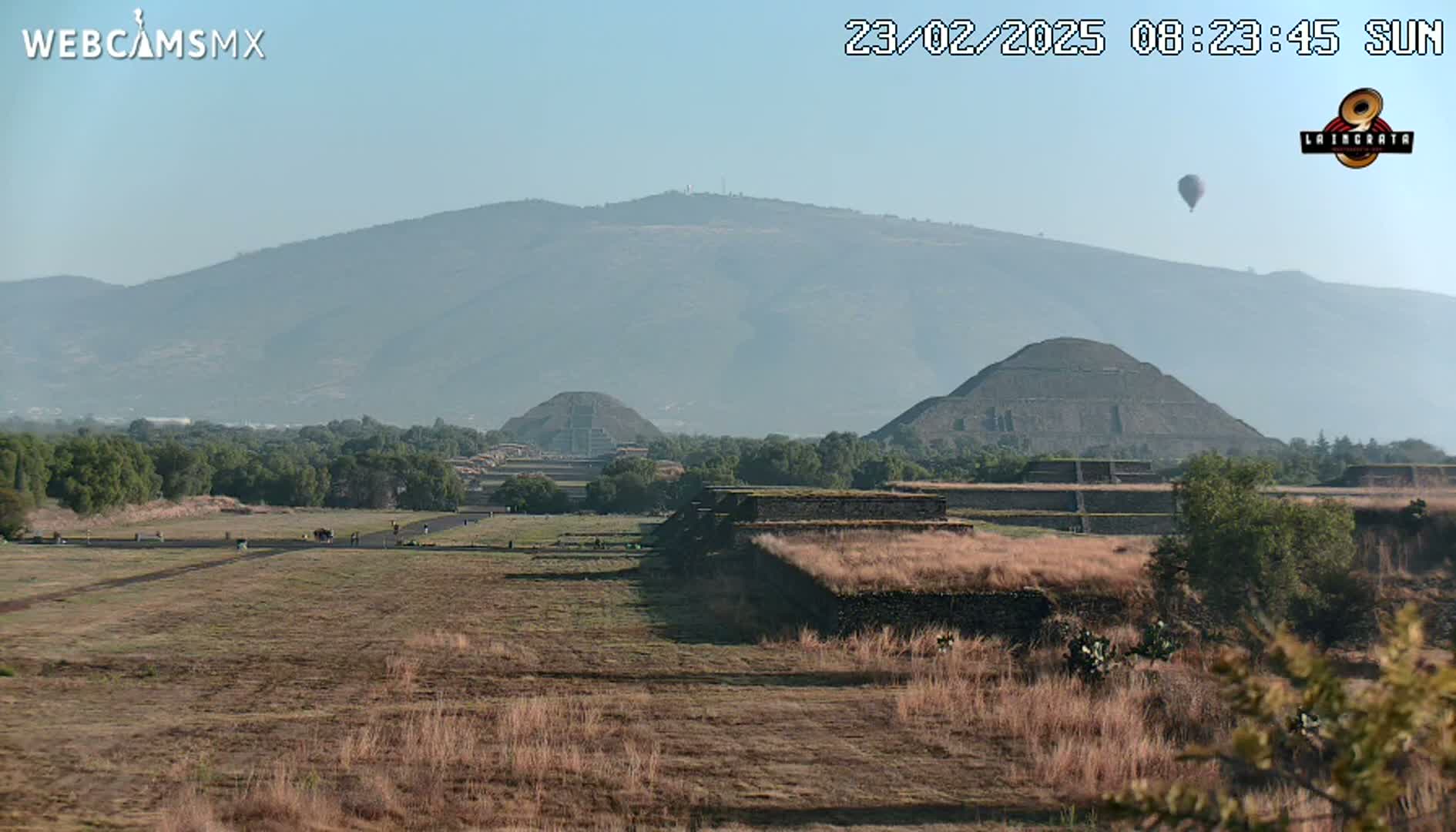 A hazy, sunny day reveals ancient stone pyramids and structures in a grassy field, with a distant mountain range and a hot air balloon in the sky.