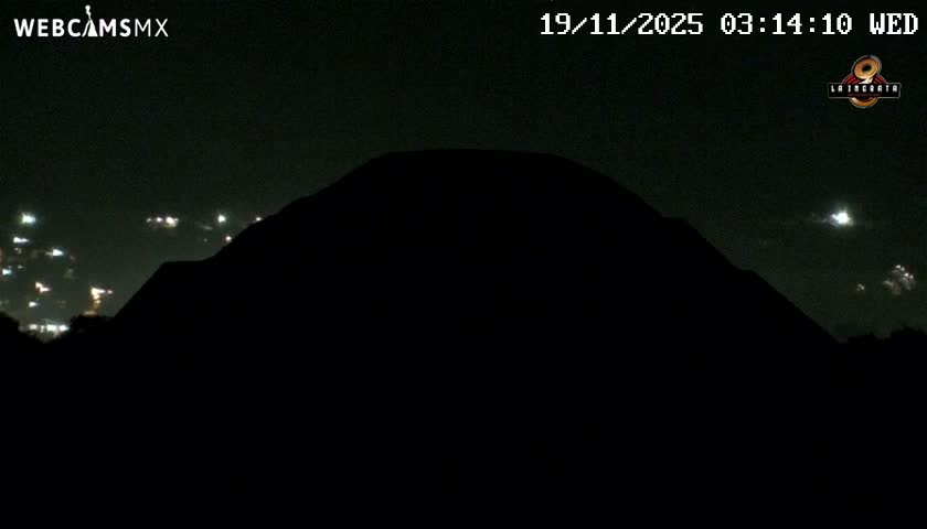 A large, dark mountain or volcano is silhouetted against a clear night sky, with distant city lights visible on the left and a bright, illuminated plume or cloud glowing on the right.