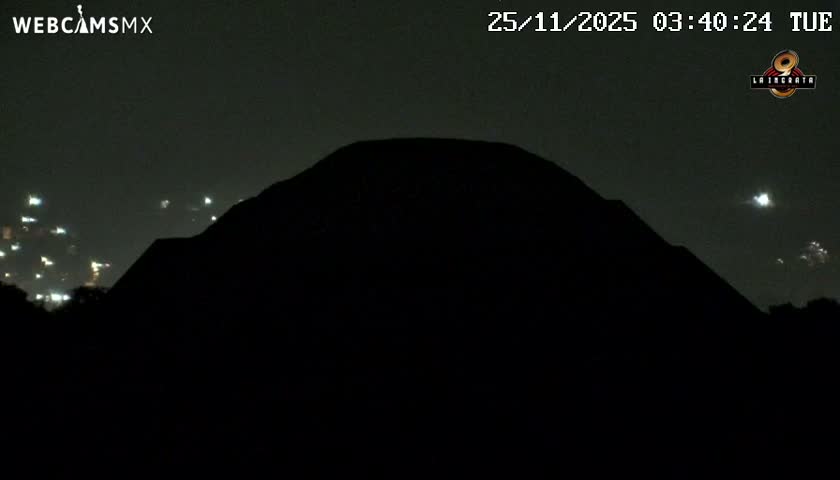 A large, dark silhouette of a pyramid dominates the foreground under a clear night sky, flanked by the scattered lights of a distant city.