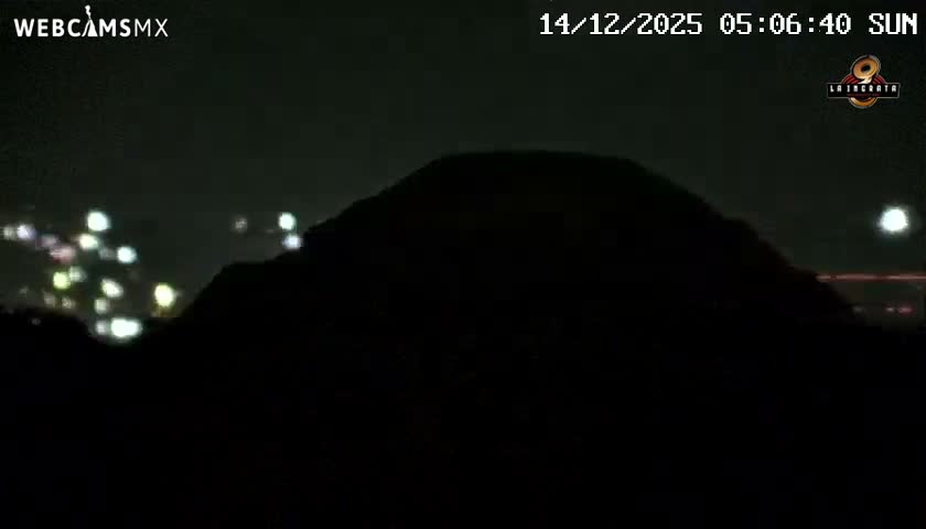 A large, dark silhouette of a pyramid dominates the foreground under a clear night sky, flanked by the scattered lights of a distant city.