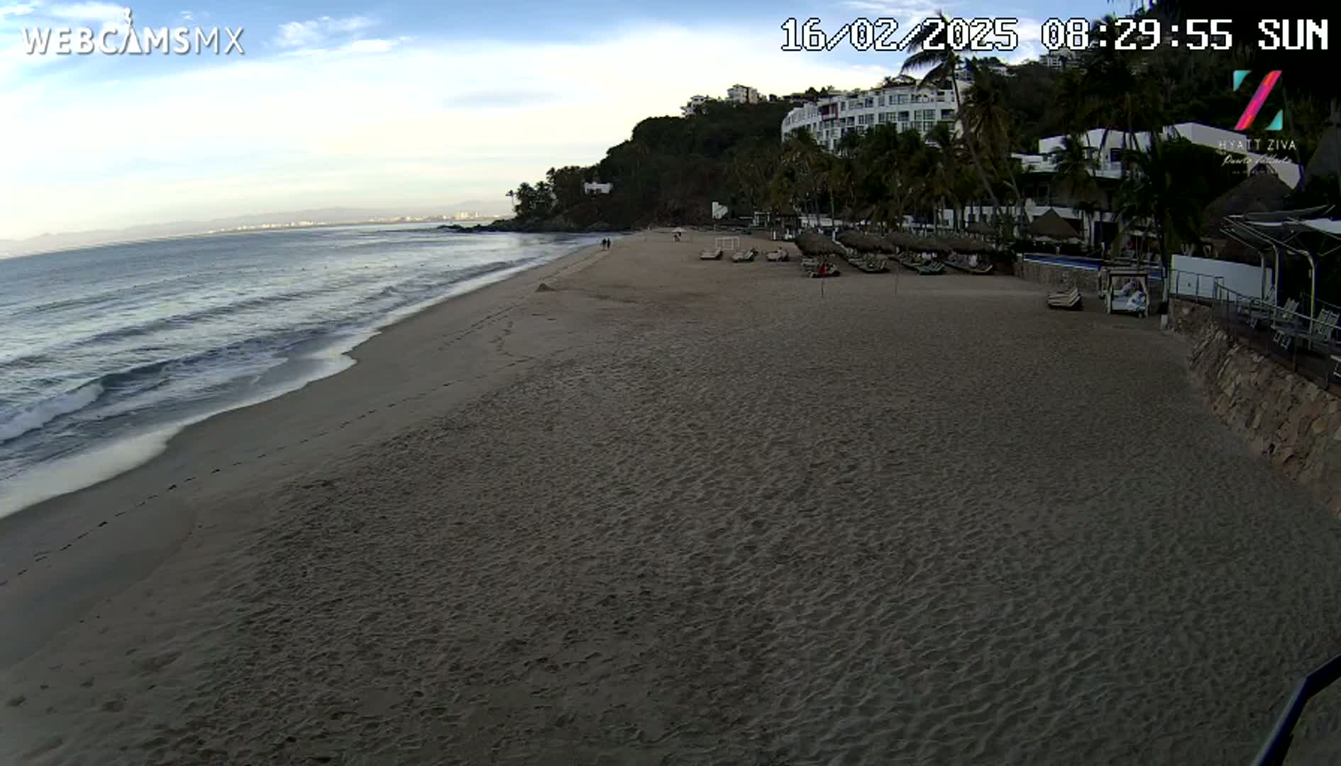 A sandy beach with gentle waves under a mostly clear sky shows a resort with palm trees and lounge chairs on the shore.
