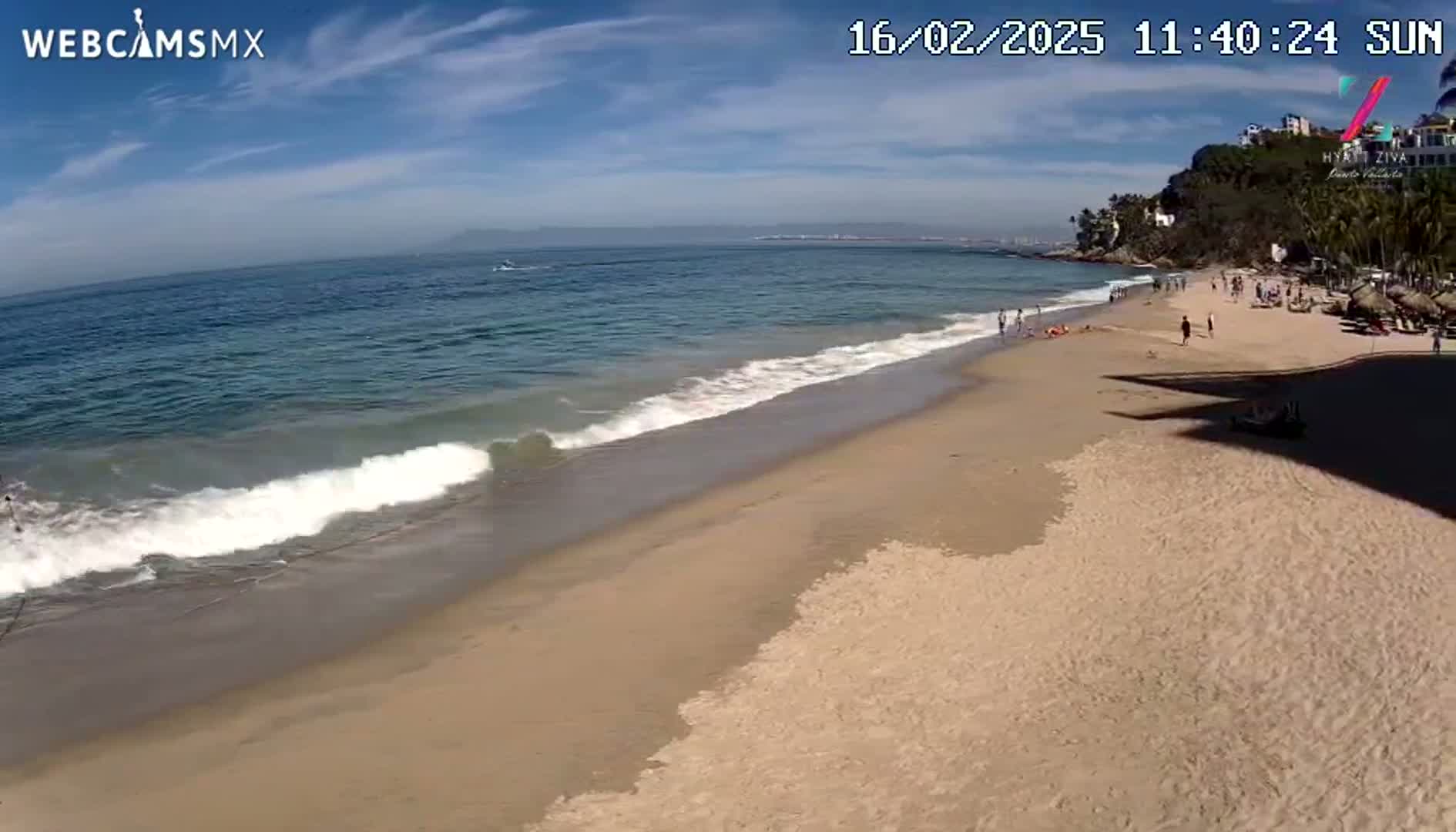 A sandy beach with gentle waves rolling onto the shore under a sunny sky, with a few people visible on the beach and in the water.
