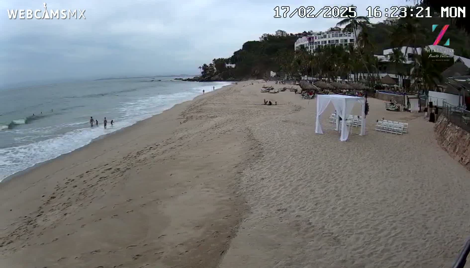 A sandy beach with a few people, ocean waves, and a white wedding setup is visible under an overcast sky.
