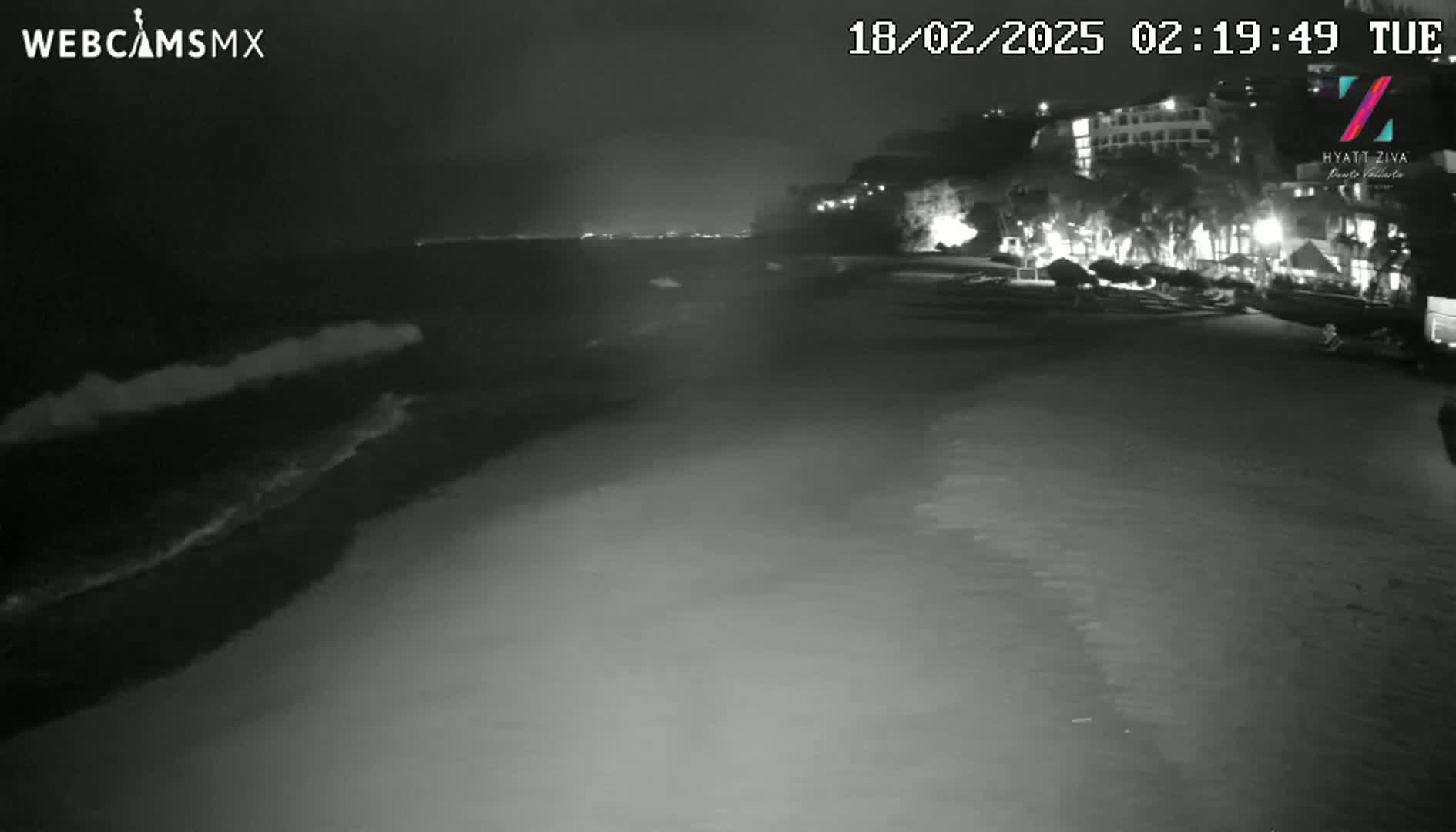 A nighttime beach scene shows dark waves lapping the shore near a dimly lit building complex.