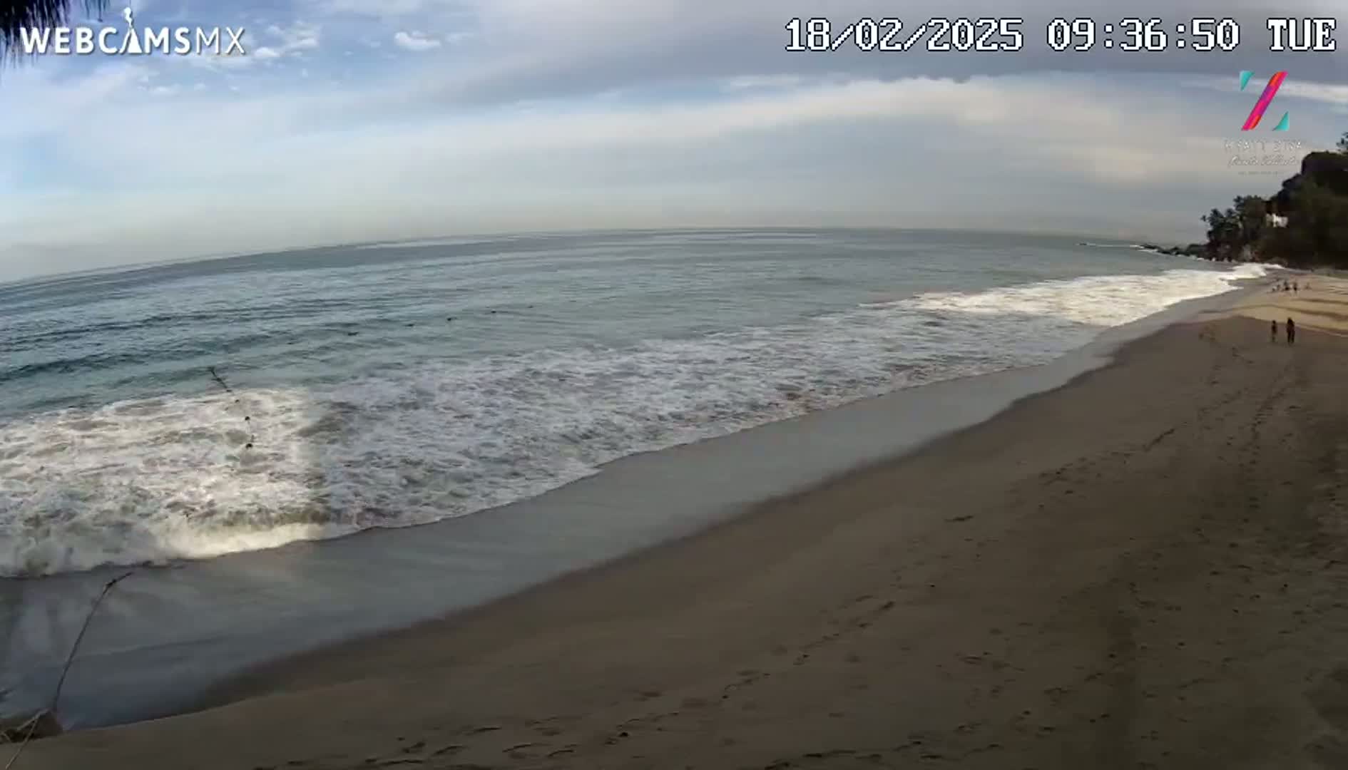 A sandy beach slopes gently down to a calm ocean with small waves under a partly cloudy sky.