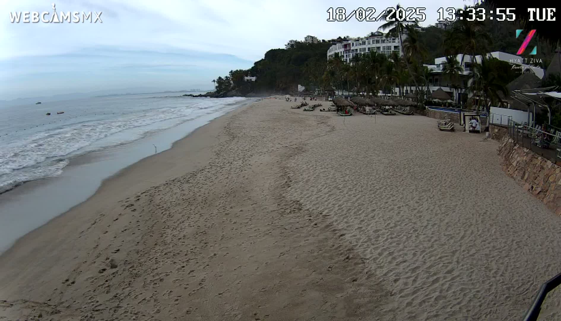 A sandy beach with gentle waves under an overcast sky, featuring a resort and palm trees on the adjacent hillside.