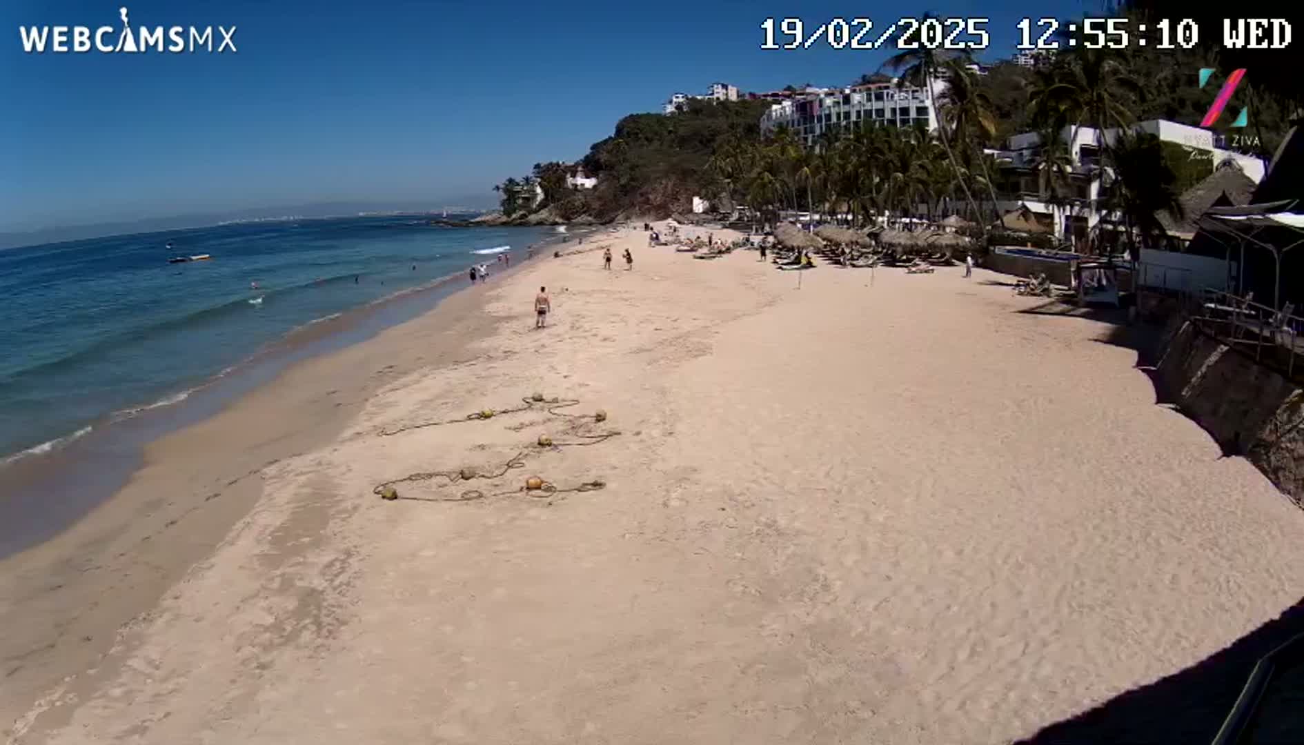 A sandy beach with a few people on it, next to a calm ocean under a clear blue sky, with resort buildings visible on a hillside.