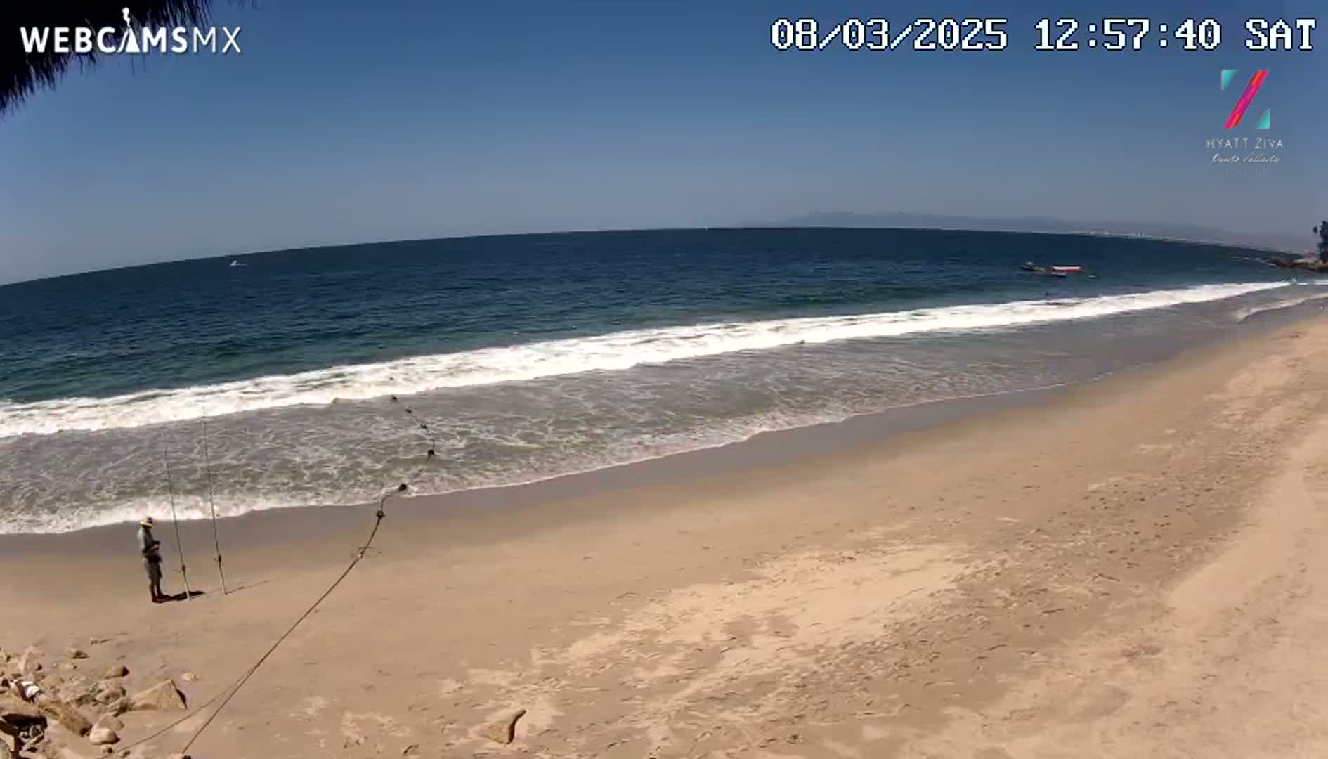 A person is fishing on a sandy beach with gentle waves under a clear blue sky.